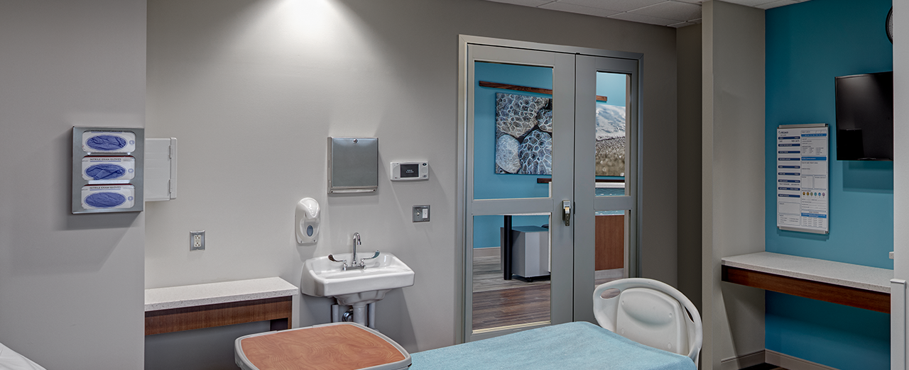 Empty hospital bed in a sterile hospital room with doors leading to hallway in background.